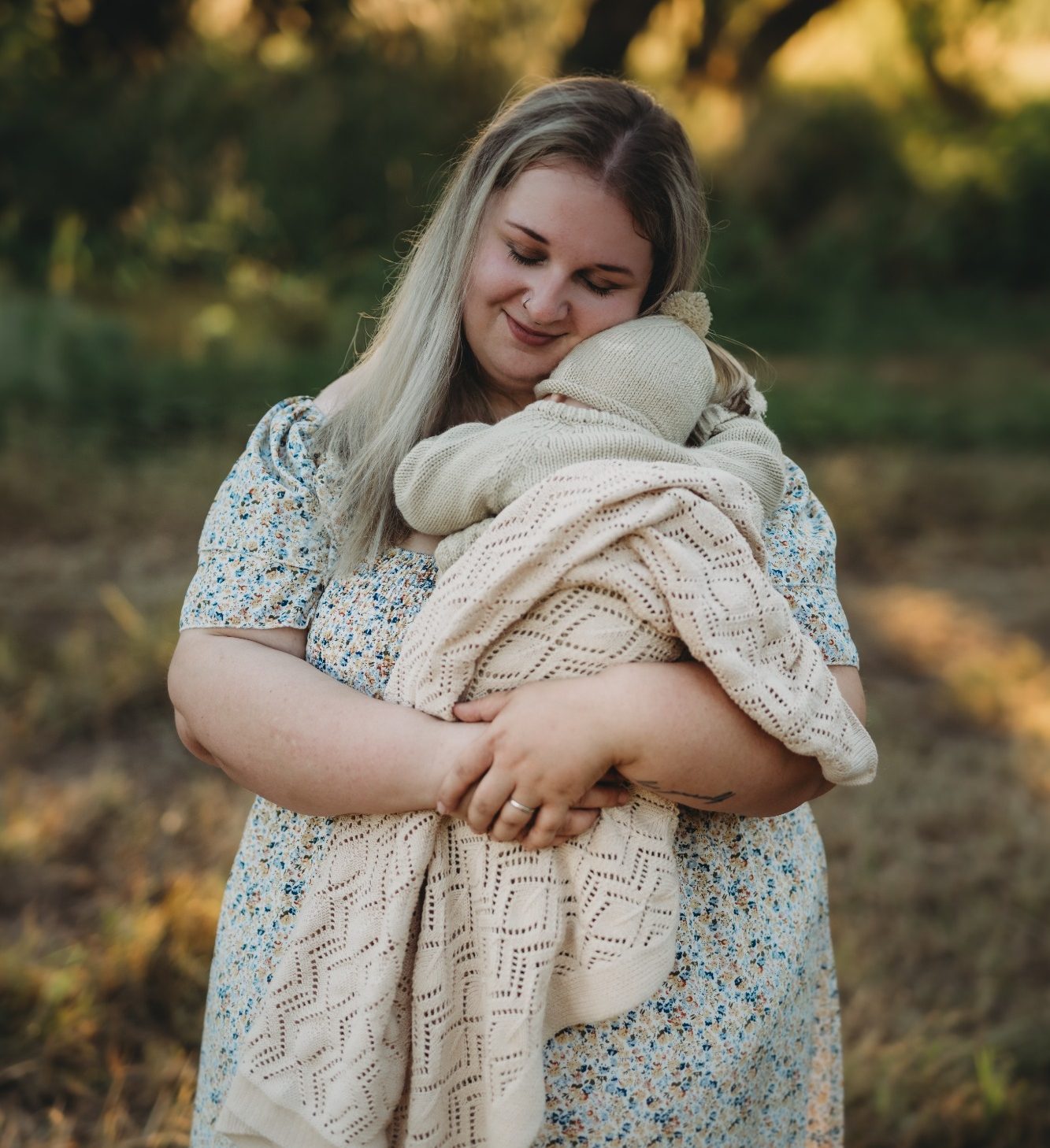 Eine lächelnde Frau hält ein Baby in einer Decke, umgeben von Natur.